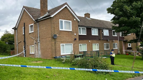 A two-storey brick-built house with police tape around it and a police officer standing watch outside