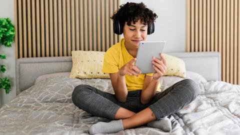 A young person wearing jeans and a yellow t shirt uses a tablet with headphones on while sitting cross-legged on a bed with grey sheets on