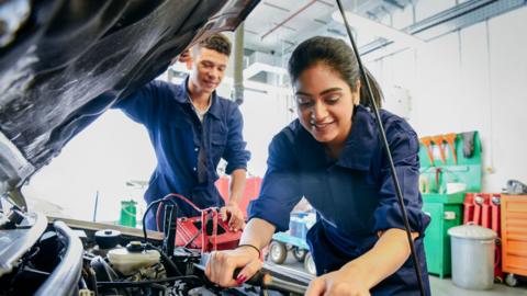 A young woman with long brown hair and a young man with short brown hair look under the bumper of a car. They are both wearing blue overalls and are standing in a workshop.