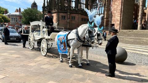 A horse-driven-hearse standing in front of Coventry Cathedral. 