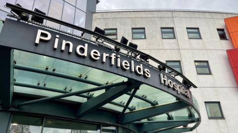 The image shows the exterior entrance of Pinderfields Hospital. A large glass canopy extends over the doorway, supported by metal framework. Mounted above the canopy is the hospital’s name in bold lettering. Behind it, a curved section of the building with multiple windows is visible.