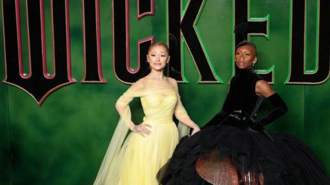 Ariana Grande wearing a yellow dress stands and smiles at the camera next to Cynthia Erivo who wears a puffy black dress. They stand in front of a green backdrop with the words 'Wicked'.