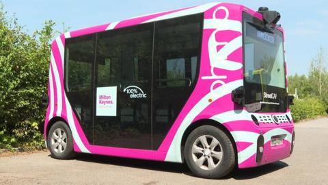 A driverless miniature bus with pink and white livery parked in a car park.