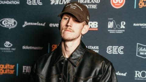 Nathan Evans standing at a red carpet event in a black leather jacket and a brown cap