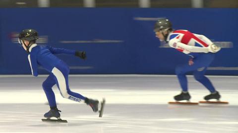 A boy and a man skate on ice they are wearing black helmets and blue and white body suits. The man's suit has a Union Jack on it.