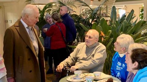 The King wearing a long brown coat while talking to four people who are sitting down. There are tea cups on the table.