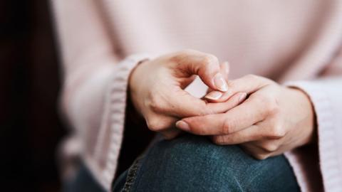 A stock image of a woman sitting on a sofa and feeling anxious. You can just see her hands, resting on her blue jeans. She is wearing a pink top. The rest of her body is blurred. 