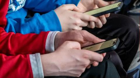Close up of 2 school pupils' hands using smart phones, one is wearing a red jumper, the other is wearing a blue jumper