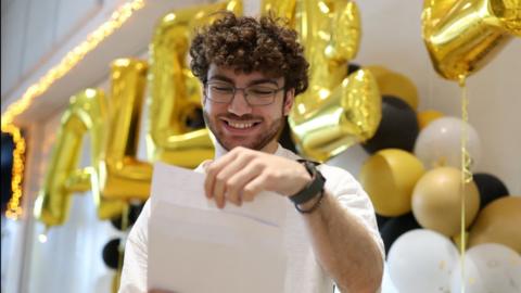A young man with glasses and brown curly hair opens an envelope to get his A-level results.