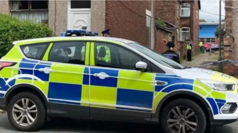 A police car in front of cordoned off alleyway with police officers visible behind it
