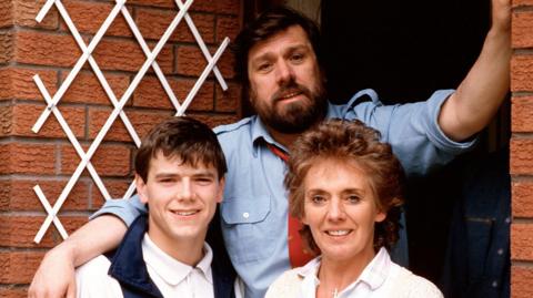 Bobby, portrayed by Ricky Tomlinson, and Sheila Grant, played by Sue Johnston are standing in front of their red-bricked home on Brookside Close with one of their children. They are all smiling at the camera in this posed photograph. 