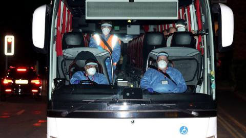 The front of a coach carrying 20 Britons to Arrowe Park Hospital. Sitting at the front are three people wearing personal protective equipment including face shields, face masks, gloves, and coveralls. The tops of some passengers heads wearing face masks can be seen.