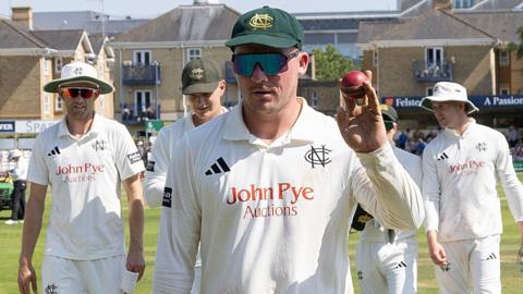 Liam Patterson-White holds up a red cricket ball with players surrounding him