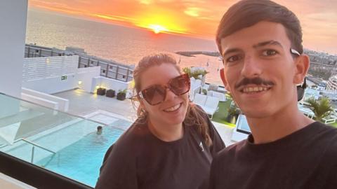 Caprice Betts and her partner Arron Sanchez Garcia standing on a balcony with a swimming pool beneath them and a view out to sea and a setting sun. She is on the left wearing a black T-shirt, has dark brown hair pulled back in a pony tail and is wearing dark glasses. He is on the right with black short hair, a black moustache and is wearing a black T-shirt. They are both smiling. 