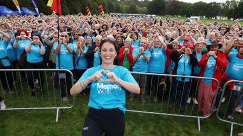 A smiling Vicky McClure wearing a blue Memory Walk shirt making a heart gesture in front of a crowd dressed in similar shirts and forming hearts with their hands