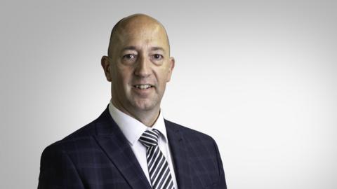 Paul Vane pictured in a black check suit. Black and white stripey tie and white shirt. He is smiling. He is middle aged with a bald head. He's being photographed in a studio, with a blank background.