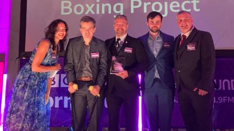 Four members of Parky Blinders, men wearing suits, collecting their award on stage.