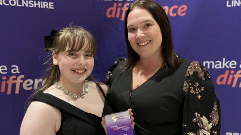 A young girl is on the left, and her mum is standing on her right. The young girl (Summer) has long brown hair that is half-up, half-down, a box fringe, and is wearing a black v-neck dress with a chunky silver necklace. She is smiling and a black bow can just be seen in her hair. She is holding a glass trophy with 'Make A Difference' written on it. Her Mumis wearing a black long-sleeved top with gold detail on the arms. She also has dark hair, worn down, just above her shoulders. She is also smiling. The backdrop (though blurred) is purple with 'Make A Difference' printed on it. 