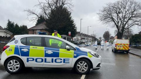 A police car is in the foreground near police tape going across the road. Behind the tape is another police vehicle and three people wearing all white are near it.