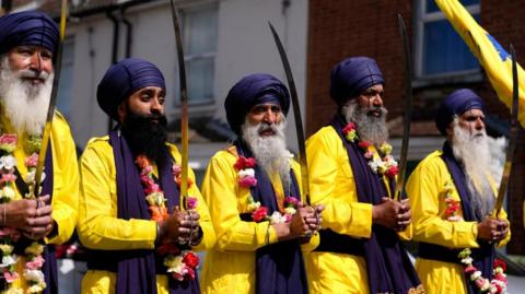 Five Sikh men in navy blue turbans and bright yellow clothes hold swords aloft as part of Vaisakhi celebrations. They have flower garlands around their necks.