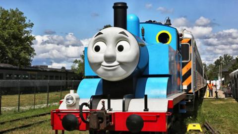 A replica of Thomas the Tank Engine. It is a blue model steam train with a grey smiley face on the front.
