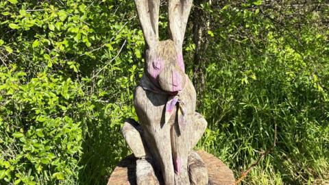 A wooden hare with pink paint splashed on its eyes, nose and paws.