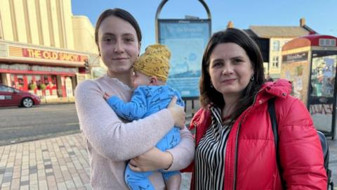 A woman in a pale pink jumper holds a baby in a blue onesie with a yellow hat. She stands next to a woman in a black and white striped shirt and a red puffer jacket. 