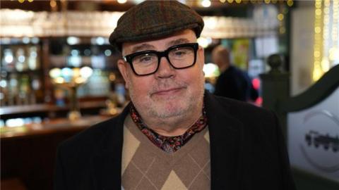 Cliff Parisi in a tartan hat and black-framed glasses standing in front of a bar in a pub. He has stubble and is wearing a flat-cap. He appears to be wearing a black coat or suit jacket.