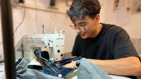 Cosmin Diaconu sits at a sewing machine repairing a pair of denim jeans. He is wearing a black T-shirt and has glasses on and black hair. 