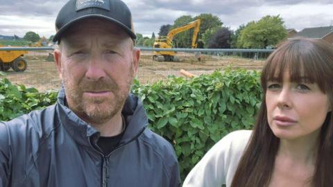 A man with a beard wearing a grey zip-up jacket and a black baseball cap. He is stood next to a woman with long brown hair and a fringe. They both have a serious expression. They are stood in their garden which has a construction site behind it with two yellow diggers on the site.