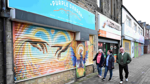 Gift Shop Purple Dragon's shutters are down. They are painted in yellow, orange and blue and who two hands reaching out to one another. Two women in jeans and dark coats are walking past and looking at the artwork. Lee Ferry is walking next to them. His hands are in the pockets of his khaki bomber jacket. He has short white hair, a beard, black circular glasses and black jeans. 