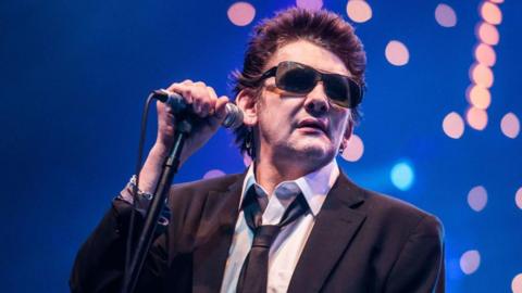 Shane MacGowan is pictured in tinted sunglasses and holding a microphone, while preparing to sing at a concert in 2012. The background is a dark blue stage backdrop with a bloom of spotlights.