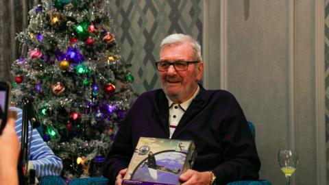 An elderly man wearing a black cardigan is holding a box containing a model Spitfire.