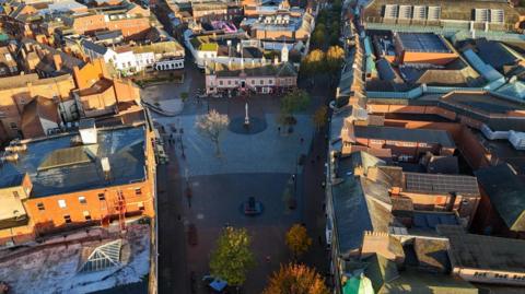 An aerial image of Market Square in Carlisle. It is surrounded by buildings, has a statue and a war memorial, trees and benches.