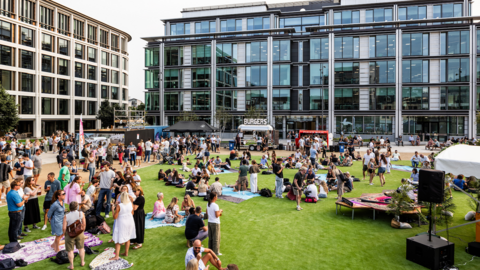 A large crowd gathers on a lawn outside modern office buildings, with people sitting on blankets, socialising, and buying food from street vendors set up in the courtyard.