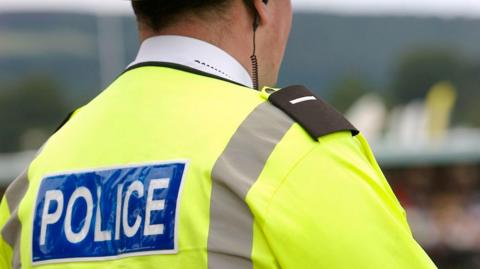 A police officer pictured from behind. They are wearing a i vid jacket with 'Police' written on it. There is a black earpiece going into their right ear.