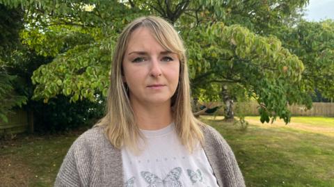 Crystal pictured looking into the camera with a serious expression. She's wearing a beige tshirt with a black illustration of a butterfly on it, and a light brown cardigan. She's stood in a garden, with a large tree behind her and grass stretching off into the distance. At the edge of the garden is a wooden picket fence, with more trees visible beyond it. 