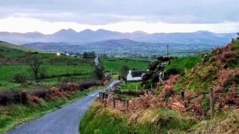 A small country road winds through green fields with mountains and grey skies behind