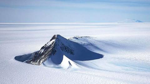 A section of the West Antarctic Ice Sheet with mountain peaks poking out of the pristine, flat, white icy surface. The sky above is light blue with light clouds.
