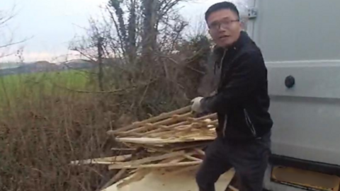 A still of bodycam footage - Chunlin Zheng is next to the back of a white van holding some wood to put on a big pile of wood next to a hedge.