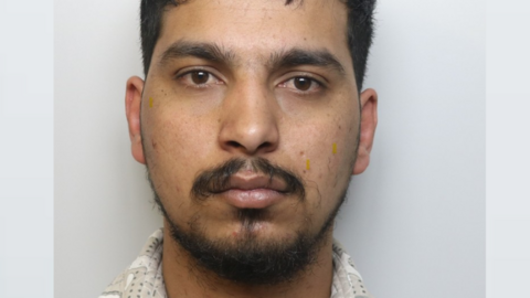 A man with a dark moustache and hair in police custody.