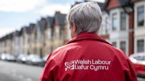 A woman wearing a red Welsh Labour jacket, with the party's logo emblazoned - Welsh Labour, Llafur Cymru - in white. The woman has grey hair and can be seen walking up a road with terraced housing, which are out of focus. Her back is to the camera.