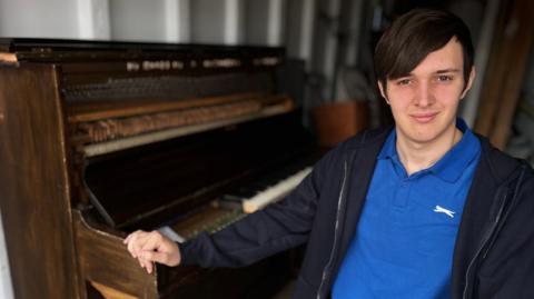Jamie is leaning on the wooden piano. He is wearing a blue polo shirt with a navy blue hooded jumper over the top. He has dark brown hair, with his fringe swept to one side. He is looking at the camera.