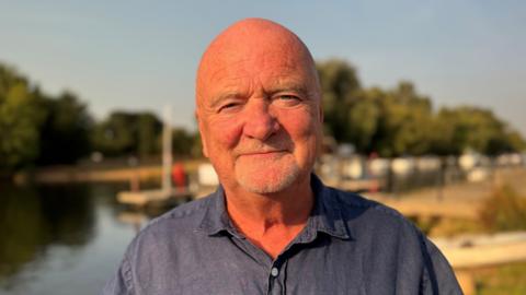 Martin Stewart smiles for the camera. He has a grey stubble beard and is wearing a dark blue open-collar shirt