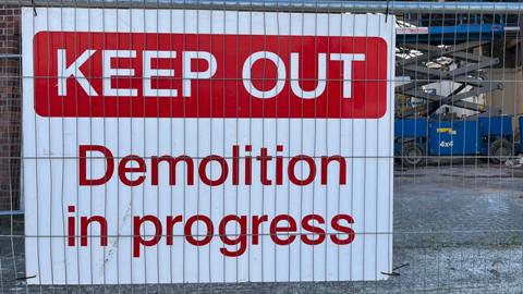 A large square sign on metal fencing that says Keep Out demolition in progress in red text on a white background