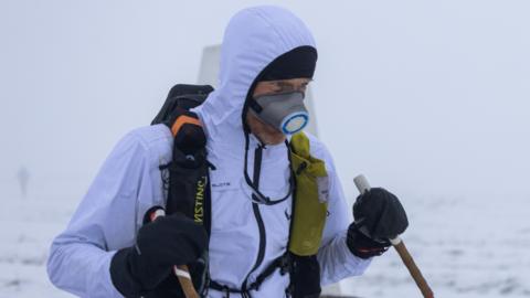 A man in a white waterproof jacket with the hood up, standing in a snowy landscape. He is also wearing a backpack and a mask over his face, and is carrying walking poles.