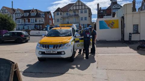Three members of HM Coastguard stand by a yellow and white marked vehicle on Westgate beach on a sunny day. Behind them are several seafront buildings and two more parked cars.