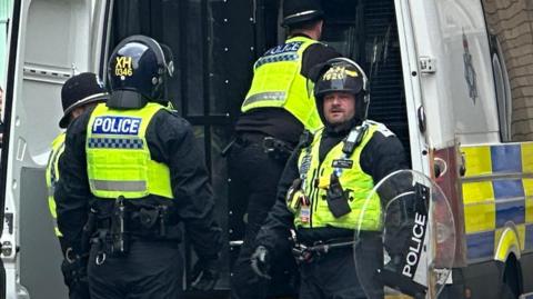 Four police officers in riot gear, including yellow hi-vis vests and helmets, at the rear of a police van. One holds a round riot shield