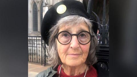 Gaie Delap in a black hat, offset to one side, with thick, round, black-rimmed spectacles. She is standing in front of a church building.