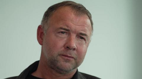 A man, who is slightly frowning, looks to the right of the camera. He has thin hair with some grey in it. He wears a denim jacket with a black T-shirt underneath.